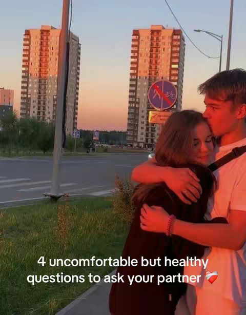 A couple in an embrace against a backdrop of two modern high-rise buildings bathed in the warm glow of a setting sun. The man is kissing the woman's head. The foreground features a patch of green grass. The sky transitions through shades of orange and pink.