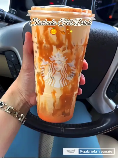 A hand holding a Starbucks drink in a clear plastic cup, inside of a car. The drink has layered colors of caramel and what appears to be the fall drink. The Starbucks logo is visible. The hand is wearing a watch with a silver band.