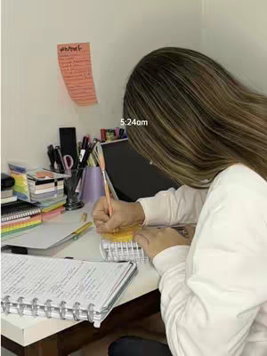 A cover image showing a woman sitting at a desk, writing on a notebook. She is seen from the back, with her head and hair partially obscuring her face. The desk holds various items, including a laptop, pens, and notebooks, suggesting a study or work environment. A post-it note is affixed to the wall.