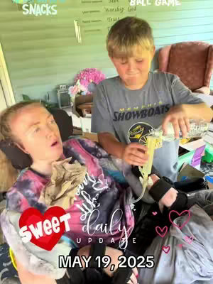 A woman in a tie-dye top is in a wheelchair, with a young boy pouring liquid into a tube connected to her. A red heart graphic and the words "SWEET DAILY UPDATE" overlay the image, along with a date. The background has a light green wall and some decorations.