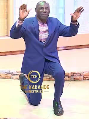 A man in a blue suit kneels with hands raised in a gesture of supplication or prayer. The background appears to be a brightly lit interior, suggesting a studio or stage setting. Overlayed on the image is a logo for The Kakande Ministries.