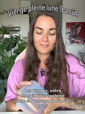 A woman with long brown hair is centered in the frame, holding a deck of tarot cards. Text overlays at the top say "Vierge pleine lune 9 août", and at the bottom says "Les vierges, votre énergie pour la pleine lune en verseau du 9 août 2025". She wears a pink shirt and has a microphone clipped to her shirt collar.