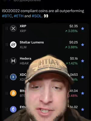 A selfie of a person wearing a camouflage hat and a dark-colored shirt, positioned in front of a phone screen showing cryptocurrency data. The phone displays a list of different cryptocurrencies, including XRP, Stellar Lumens, Hedera, XDC, Bitcoin, and Ethereum, along with their current prices and percentage changes. The background has a dark theme. The person is looking at the camera and speaking.