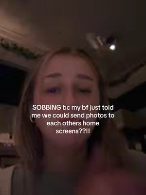 A selfie-style shot of a young woman with blonde hair, looking towards the camera with a slightly distressed expression. The background is softly lit, with visible architectural details like a ceiling fan and an indistinct screen. Text overlays include "SOBBING bc my bf just told me we could send photos to each others home screens??!!"