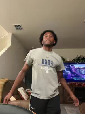 A man with dark, curly hair stands in a living room, arms outstretched. He wears a gray t-shirt with a logo and black shorts. Behind him, a TV displays a video, and a sofa with decorative pillows is visible. The lighting suggests an indoor setting.