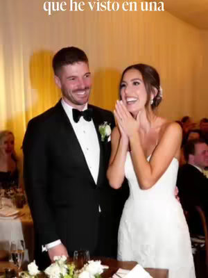 A candid shot of a newlywed couple celebrating their wedding. The groom is smiling, wearing a black suit, bow tie, and boutonniere, while the bride is beaming with her hands clasped near her face, wearing a white strapless wedding dress. They are positioned in front of a background with warm lighting and wedding guests.