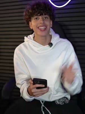 A young man with curly hair is smiling while holding a black smartphone. He's wearing a white hoodie and dark pants. The background is dark, showing the horizontal blinds.