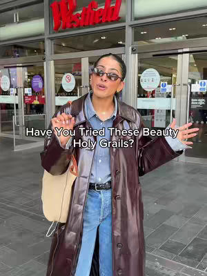 A woman stands outside a Westfield shopping center, wearing sunglasses, a long brown coat, and jeans. The text overlay says 'Have You Tried These Beauty Holy Grails?' The image has a modern and clean composition, conveying a sense of fashion and lifestyle.