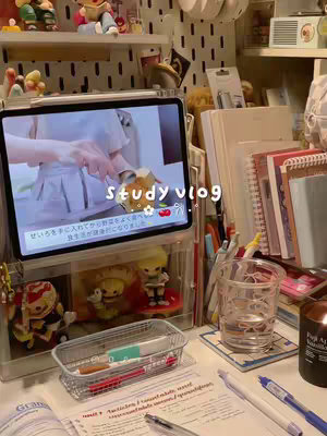 The image shows a desk setup with a tablet displaying a video, likely of someone preparing food, within a clear acrylic organizer. Around the tablet are study materials: open books, pens, and a clear glass with water next to a lit candle. The desk is made of a light colored wood and is cluttered with books, stationary, and decorations creating a cozy study environment. The overall color palette is warm with whites, browns, greys, and touches of clear or transparent materials.