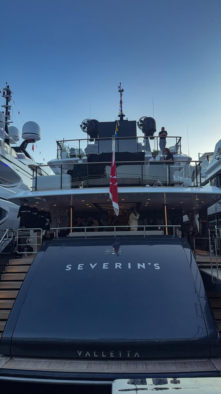 The image shows the stern of a large yacht with the name "Severin's" and the city name "Valletta" in white letters. A red and white flag is displayed in the center. Several people are visible on the yacht's upper decks, and the background is the blue sky. Other yachts are visible on either side of the featured yacht, creating a scene of luxury and elegance.