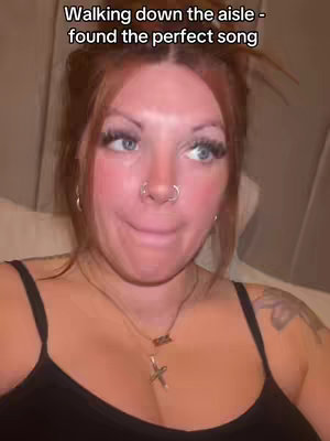 A selfie-style shot of a woman from the chest up. She has fair skin, red hair pulled back, blue eyes, and is wearing a black tank top and a necklace. The background is softly blurred. Text overlay at the top of the image.