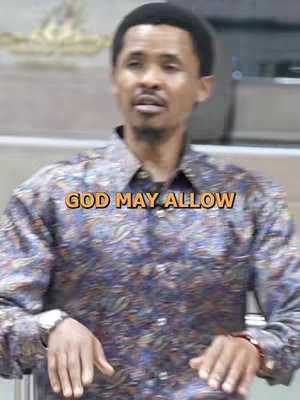 A man in a paisley collared shirt is the main focus of the image, giving a direct gaze upward. The text overlay, "GOD MAY ALLOW," is placed across the center of the image. The background appears blurred, but suggests an indoor setting.
