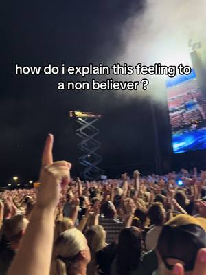 The cover image shows a large crowd at an outdoor event, likely a concert or festival, with the sky above. Many people are raising their hands in the air, and some are holding up phones to record. The foreground features a person's upward-pointing finger, which is highlighted. A text overlay across the top asks: 'how do I explain this feeling to a non believer?' In the background, there is a large stage with screens, and a tall, angular structure rises in the background. The lighting seems to be a mix of stage lights and natural light with a dark and cloudy atmosphere.