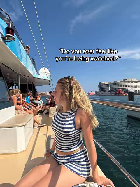 A young woman with blonde hair, wearing a blue and white striped outfit, sits on the edge of a boat. She is turned to her left, with a calm, inquisitive expression. The background features the ocean, a city skyline, and a blue sky with some clouds.