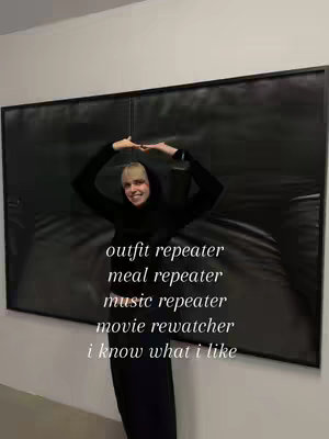 A woman in a black hoodie stands in front of a large black painting hanging on a white wall,. She is smiling and making a square with her hands. Text is overlaid on the image.
