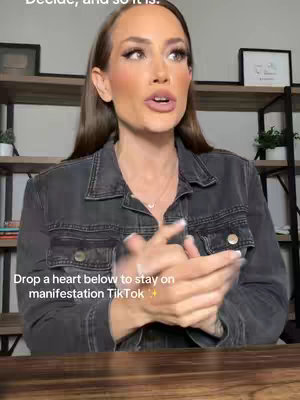 A woman with long brown hair is shown from the chest up, speaking directly to the camera. She wears a denim jacket over a white top, with natural makeup. Behind her are shelves with various objects and a framed picture on the wall. The background is softly lit, emphasizing the subject.