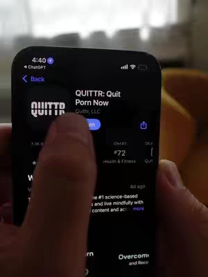 The image is a screenshot of a phone displaying the app store. The phone is held by a hand in the foreground, with the app store interface of "QUITTR: Quit Porn Now" visible. The interface showcases the app's icon, name, description, ranking, and an "open" button. The background appears to be a slightly blurred interior scene possibly with a window.