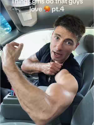 A man is seated inside a car, with the driver's side door and dashboard visible. He is flexing his arm to show off a large bicep while talking to the camera. Above him, a gray sun visor is extended. The background shows the car's interior, including a seat and console.