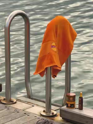 An outdoor scene at a pool. A chrome ladder for a pool leads up from the edge of a wooden deck. A bright orange towel hangs over the ladder. There is a bottle and container next to the ladder.