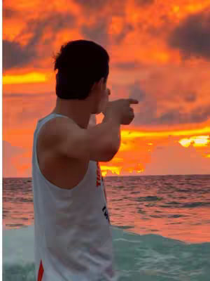 A man stands at the edge of the water, facing away from the camera and pointing his finger towards a vibrant sunset sky. The sky is filled with shades of orange and yellow, with some cloud formations. The sea is calm, reflecting the colors of the sky. The man is wearing a white tank top, and only visible from the back. The overall composition evokes a sense of peace and admiration for the scenery.