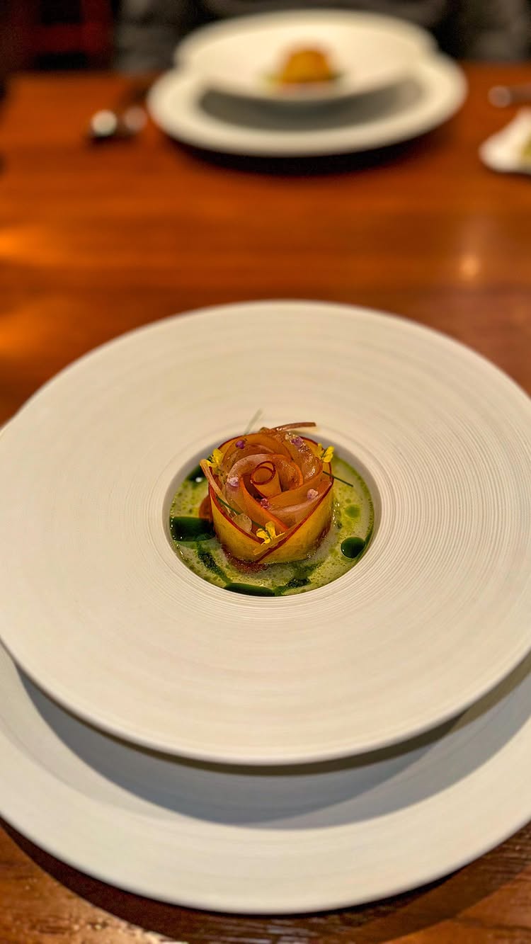 A close-up shot of a finely plated dish. The main element is a flower-shaped arrangement, composed of thinly sliced, light-brown fruit with a darker edge, possibly an apple. It sits atop a light green sauce, around which are small green dots and flower petals. The dish is presented in a white, round plate, and sits on another, larger white plate. The background is a dark, wooden surface, likely a table.