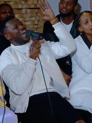 The cover image captures a live performance. The main subject, a Black man with short, dark hair, is singing into a microphone, his mouth wide open. He wears a white jacket over a white shirt and has a gold wristwatch. Behind him, several blurred figures are visible, also appearing to participate in the performance. The background includes a wooden wall, and the overall lighting suggests a well-lit indoor setting.