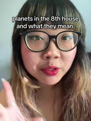 A woman with long, light brown hair and glasses is centered in the frame of a selfie-style video. She's wearing glasses with thick black frames and her skin is fair. The framing is a close up shot of the face, the text 'planets in the 8th house and what they mean' appears as an overlay which has a black background with white text.  She is looking directly at the camera.