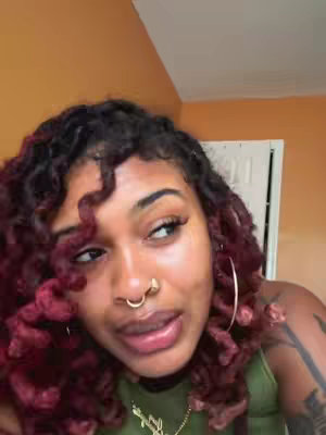 A person with dark curly hair dyed with red, wearing a green top and a nose ring, is looking to the side in a close-up shot.