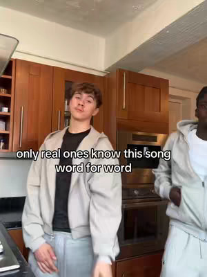 Two young men are positioned in front of wooden kitchen cabinets and appliances. One man, the primary subject, is in the center, wearing a light-colored jacket over a black shirt; he looks towards the camera. The other man, on the right, is partially visible and wearing a matching jacket. A text overlay at the bottom reads, 'only real ones know this song word for word'. The lighting appears natural or from overhead sources.