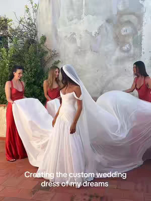 A bride in a white wedding gown is standing as three women, dressed in red dresses, hold up and arrange her long train and veil. The scene is set outdoors on a terrace, with a textured wall as a backdrop and greenery visible. The bride's off-the-shoulder gown and veil form a dramatic silhouette against the composition.