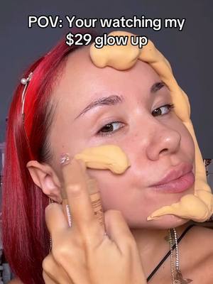 A close-up shot of a person's face with makeup applied. The person is looking towards the viewer with what appears to be makeup on their face and hands. Overlaid text at the top reads "POV: Your watching my $29 glow up". The background is a muted tone of various colors.