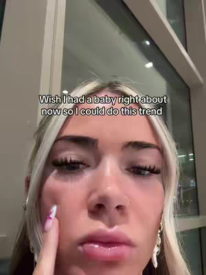 A close-up shot of a young woman's face, with blonde hair, visible eyelashes, and a small nose ring. She appears to be touching her cheek with a manicured fingernail, and the background is out of focus.