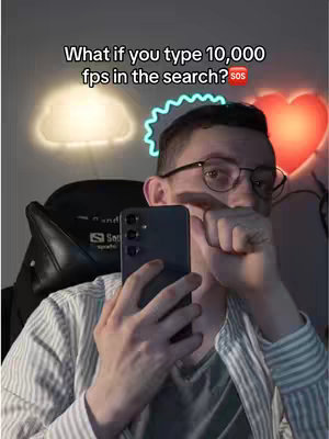 A man with glasses holds a black phone in front of his face. The background features a soft, illuminated cloud-shaped light on the left and a glowing heart-shaped light on the right. The man's hand covers his mouth in a surprised or secretive gesture. He is wearing a striped shirt and sitting in a gaming chair, suggesting a tech-related or gaming environment.