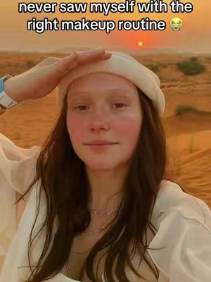 A woman in a white beanie and top poses in a desert landscape with a hand over her forehead. The background features a blurred desert scene with the sun setting or rising. Overlayed text at the top reads 'never saw myself with the right makeup routine'.
