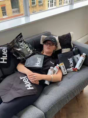 A woman is lying on a gray couch, surrounded by various Huel products. She is wearing a black cap, glasses, and a black t-shirt with the Huel logo. Bags and bottles of Huel are arranged around her, giving the impression of a Huel enthusiast lounging in a setting with a window behind her.
