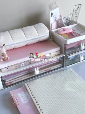A well-organized desk with light pink and white accents. There are various items like notebooks, pens, a pencil case, and a small bunny figure. The overall aesthetic is clean and tidy, typical of study setups.