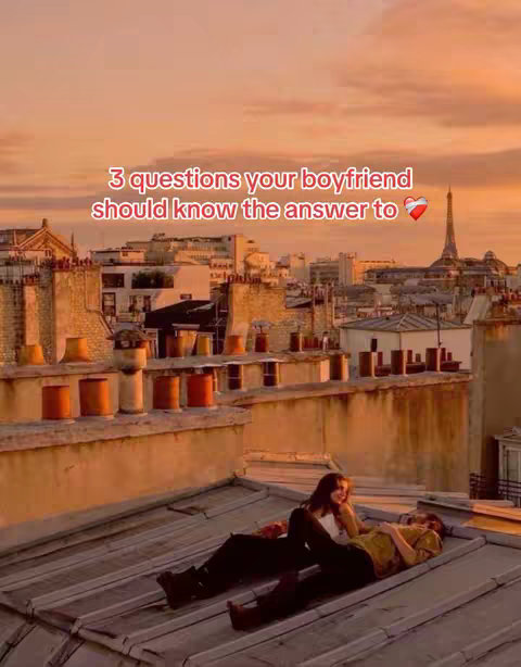 A romantic rooftop scene at sunset. A couple lies on a sloped roof with a cityscape and the Eiffel Tower in the background, clouds with orange hues fill the sky. The foreground shows the couple lying on the roof; one person is wearing a white top and the other in a golden top. The shot evokes intimacy and a sense of shared experience.