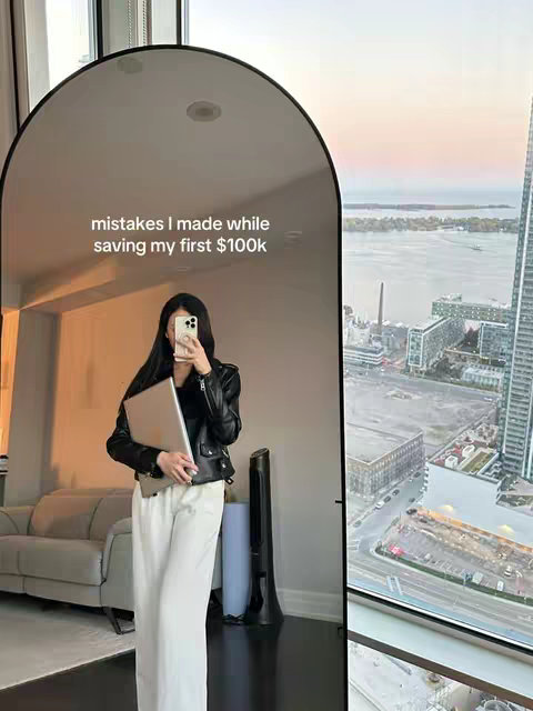 A woman in a black jacket and white pants is standing in front of a tall arch mirror, holding a laptop and her phone. The background shows a room with a couch, a fan, and a large window with a cityscape view, and the hook text is on the mirror.