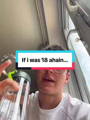 A selfie-style shot taken indoors with a man holding a water bottle, the camera is close up and angled slightly upwards. The man has short, dark hair and is wearing glasses and a white t-shirt. The background includes the edge of a window and a handle, and an overlaid banner that reads "If i was 18 ahain..."
