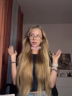 A young woman with long blonde hair and glasses stands in a room with her arms raised and hands open. The text overlay "Ready?" appears in a white block at her mouth level. Behind her, a partial view of a curtain and a bulletin board are visible.