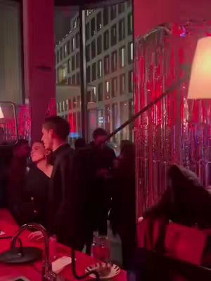 A dimly lit interior shot of what seems to be a restaurant or bar. Several people, including a man and a woman, are seen casually conversing. The background features a cityscape viewed through a large window, casting a soft glow into the interior. Red lighting adds a warm ambiance to the scene.
