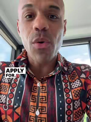 A medium shot of a man looking at the camera, with a short haircut and bald head. He is wearing a colorful patterned shirt with red, orange, and black designs. A text overlay in the corner reads "APPLY FOR."