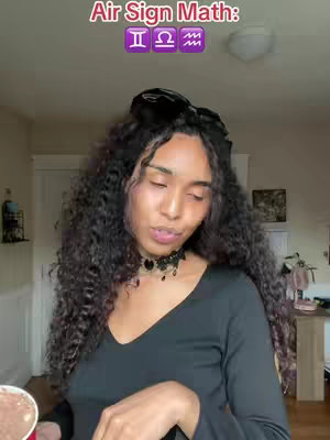 A woman with long, curly dark hair looks towards the camera. She wears a black top and a black choker. Above her head, text reads 'Air Sign Math: ' with the air sign symbols. The background is the interior of a room. The lighting is natural, illuminating the subjects.