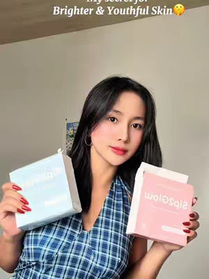 A young woman is holding two product boxes in each hand, smiling at the camera. The box in her left hand is light blue, and the box in her right hand is pink. The background seems to be a beige wall.