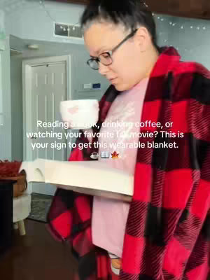 A woman wearing glasses and a red and black plaid wearable blanket, is in a cozy living room setting, holding a cup and a book. Text overlaid on the image reads, "Reading a book, drinking coffee, or watching your favorite fall movie? This is your sign to get this wearable blanket."