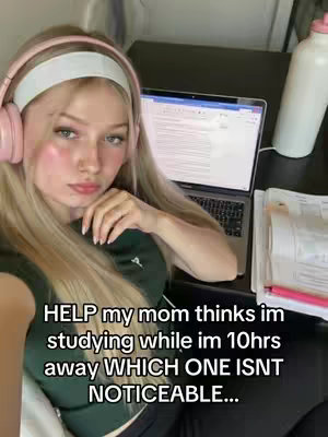 A young woman with long blonde hair and pink headphones sits at a desk with a laptop and papers. She is wearing a dark green top and has a slightly bored expression. The background includes a white wall and a glimpse of a white bottle.