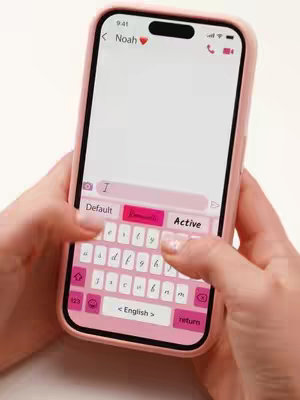 A person holding a pink iPhone with a pink keyboard theme. The screen displays a messaging app interface, and the person's hands are visible, with fingers pressing the virtual keyboard.