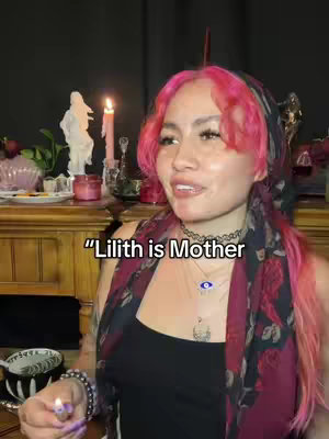 A woman with pink hair and a scarf is in front of a wooden cabinet with objects in the background. Text that says "Lilith is Mother" is overlaid on the image.