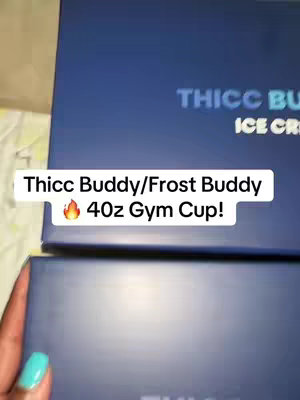 The image shows a close-up view of two dark blue boxes, likely product packaging, with text overlays. The boxes are positioned at a slight angle, with the bottom one partially visible at the bottom. A hand, adorned with a light blue painted fingernail, is visible holding up a box. The main text overlay reads, "Thicc Buddy/Frost Buddy" and "40oz Gym Cup!", with a flame icon next to it. The "Thicc Buddy" name is also visible on one of the boxes. The overall composition is simple, with a focus on product presentation within a well-lit setting.