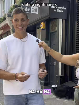 A man in a white t-shirt smiles, holding a microphone outdoors, while a person's arm with a bracelet holds the microphone. Above the man's head is a street sign, blurred. In the bottom corner is a text overlay 'HA HA HA'. The background includes a store with an Abercrombie & Fitch sign.